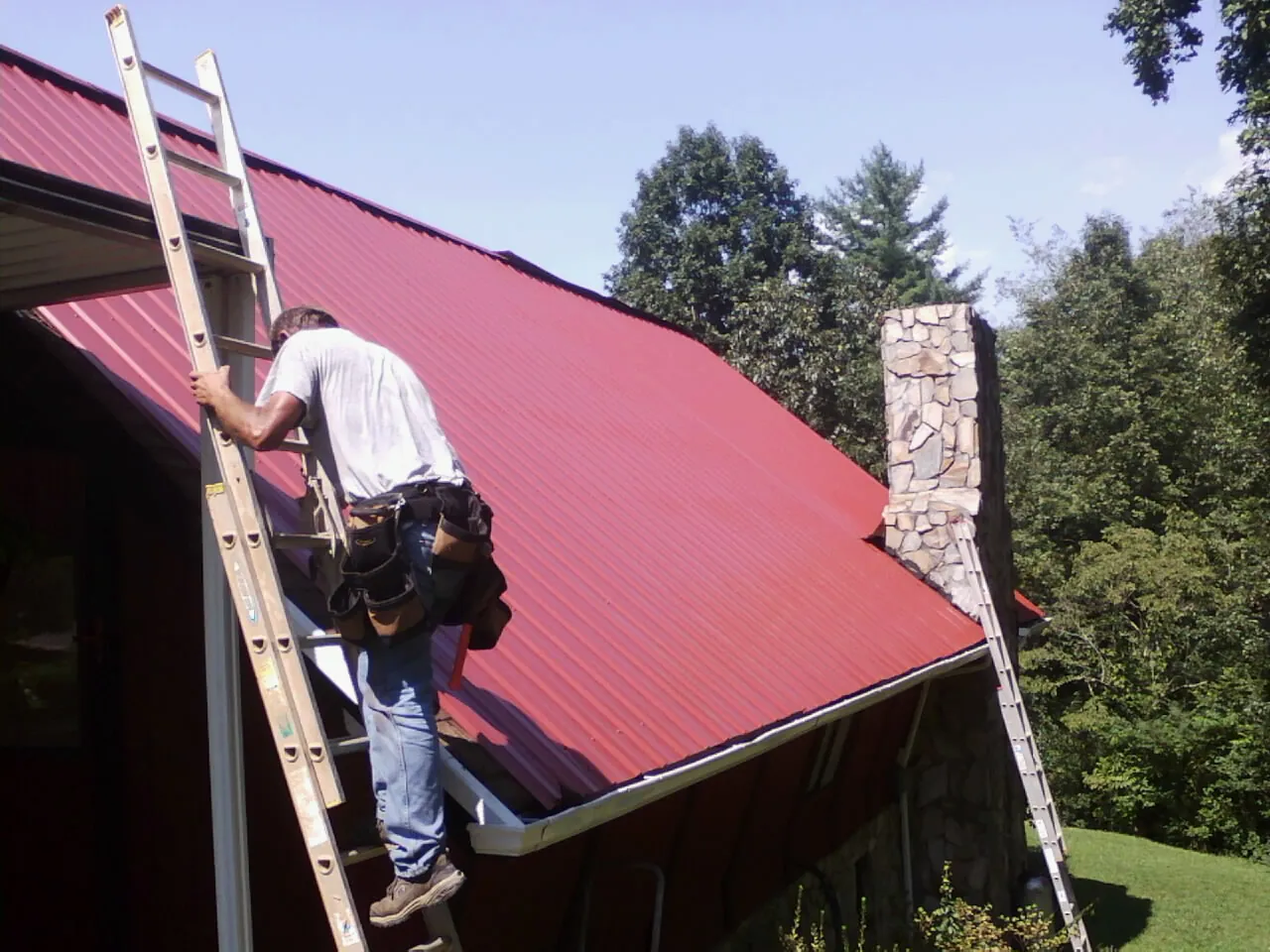Roofer on a ladder beside a red metal roof and stone chimney — Soffit & Fascia Repair by Roofing Brothers in Stirling